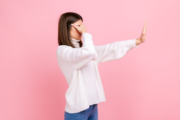 Side view portrait of female standing with outstretched arm, covering eyes with palm, stop gesture, wearing white casual style sweater. Indoor studio shot isolated on pink background.