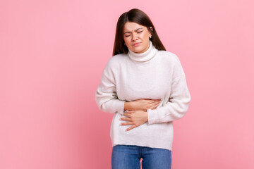 Ill brunette woman suffering acute pain, cramps from indigestion or gastritis, appendicitis symptoms, wearing white casual style sweater. Indoor studio shot isolated on pink background.