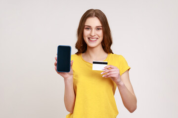 Cheerful teenager girl in yellow casual style t-shirt holding and showing empty display smartphone and credit card, satisfied with easy mobile banking. Indoor studio shot isolated on gray background.