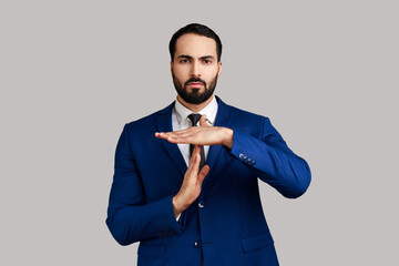 Serious strict bearded man showing time out gesture, looking with imploring eyes, hurry to meet deadline, wearing official style suit. Indoor studio shot isolated on gray background. - obrazy, fototapety, plakaty