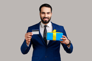 Portrait of smiling positive bearded man showing credit card and gift box, looking with amazement,...