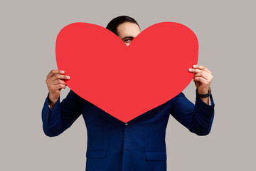 Man hiding face behind big red heart and looking at camera with curious prying eyes, expressing i love you, feeling affection fondness. Indoor studio shot isolated on gray background.