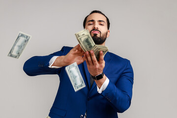 Bearded man scattering dollars with arrogant grimace, boasting wealthy life, concept of careless money spending, wearing official style suit. Indoor studio shot isolated on gray background. - obrazy, fototapety, plakaty