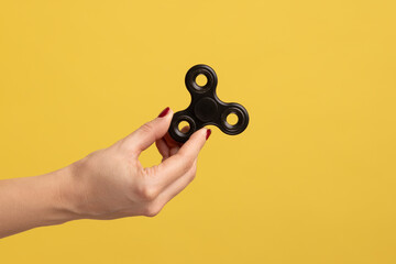 Closeup side view profile portrait of woman hand holding black fidget spinner, stress relieving toy. Indoor studio shot isolated on yellow background. - obrazy, fototapety, plakaty