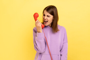 Housing maintenance call center. Angry brown haired woman shouting into retro phone handset, irritated by customer call, wearing purple hoodie. Indoor studio shot isolated on yellow background.