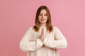 Fototapeta premium Portrait of blond woman standing with closed eyes, keeping palms together, doing yoga exercise breath technique reduce stress, wearing white sweater. Indoor studio shot isolated on pink background.