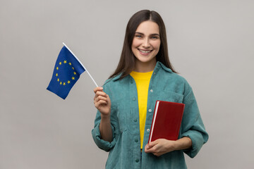Portrait of positive student woman holding books and Europe Union flag, education abroad, looking smiling at camera, wearing casual style jacket. Indoor studio shot isolated on gray background.