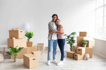 Glad young caucasian male and female in casual hugging in room with cardboard boxes, renting flat...