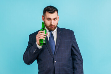 Bearded young adult drunk businessman wearing official style suit posing with bottle of alcohol in hands and having fanny facial expression. Indoor studio shot isolated on blue background.