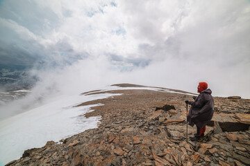 Fototapeta premium Awesome minimalist mountain landscape with hiker at very high altitude in dense low clouds. Wonderful alpine scenery with man on stone hill with snow in thick clouds. Man and majestic mountains nature