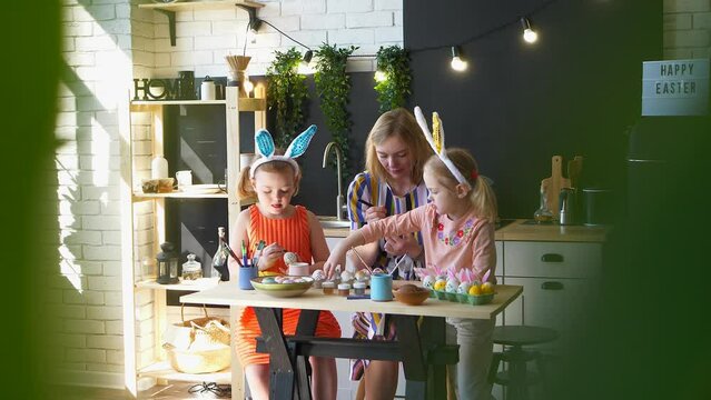 Mom and her daughters are coloring eggs for Easter holiday at home in the kitchen, mother spend her time with her children, happy Easter preparations. View trough blurred leaves.