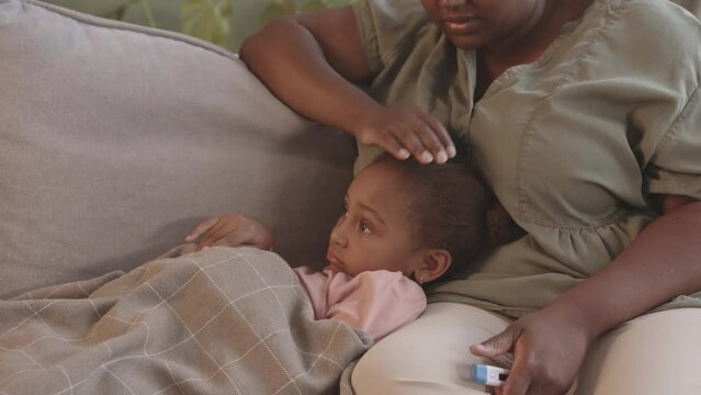 Medium slowmo of Black woman taking care of her sick 5 year old lying under plaid on sofa with thermometer