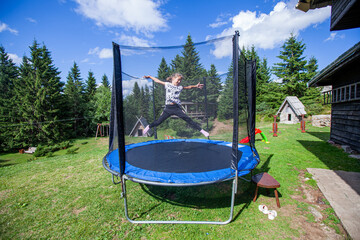 Happy little girl jump on trampoline in backyard. Child fun outdoor. Summer day. Blue sky. Nature landscape in background. - obrazy, fototapety, plakaty
