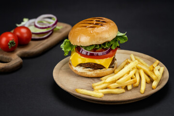 Tasty homemade beef burger with fresh ingredients served on little wooden cutting board with french fries on dark background, close-up view.