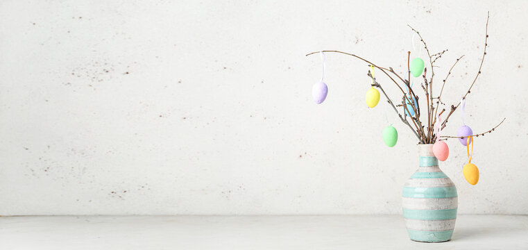 Easter eggs hanging on tree branches in vase against light background with space for text