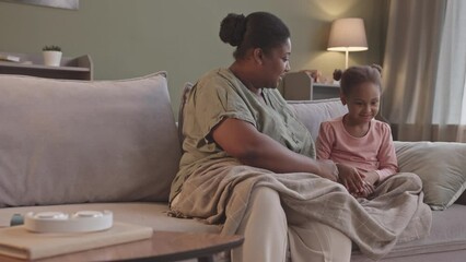 Medium slowmo of young curvy African-American woman using asthma inhaler after playing with her 5 year old daughter spending leisure time together on sofa in living room