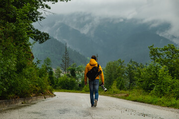 Man is walking alone down the road in misty mountains wearing backpack and with camera in his hand