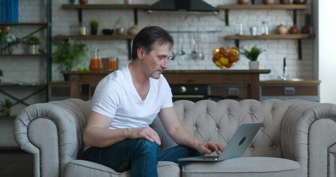 Portrait of caucasian handsome mature man sitting on sofa in living room in apartment and typing on laptop computer, working distantly at home