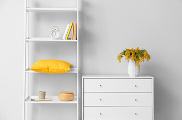 Vase with mimosa flowers on chest of drawers and shelving unit near light wall
