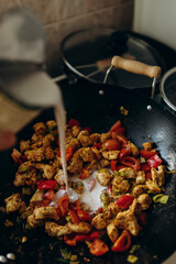 pour the coconut cream into the white meat in the wok. pouring cream on meat with vegetables in a frying pan top view. pieces of chicken fillet with spices and vegetables in a wok.