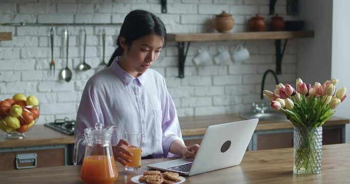 Asian woman sit at table in cozy kitchen working at laptop computer, freelancer texting e-mail response to client, telework activity from home using internet and drinking juice