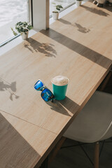 A cup of cappuccino with you and with sunglasses on the table near the window. Coffee break in a cafe on a sunny day. Stylish blue glasses and delicious coffee