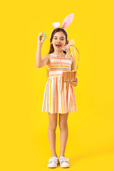 Cute little girl with bunny ears and Easter basket on yellow background