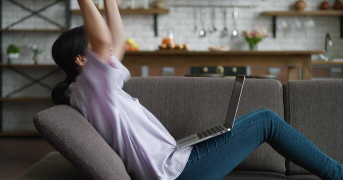 Happy young asian woman celebrate success win online bet and showing yes gesture. Lucky female freelancer reading great news online using laptop while resting on cozy sofa at home.