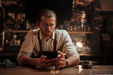 Young caucasian male bartender sitting alone at bar texting on cellular device 