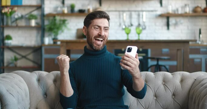 Happy businessman enjoying success win on mobile phone on sofa at home. Joyful male reading good news on smartphone, showing yes gesture. Surprised man celebrating victory on phone in living room