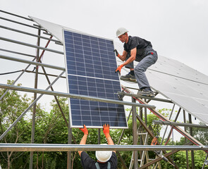 Male worker in safety construction helmet giving solar module to colleague. Two men in workwear building photovoltaic solar panel system. Concept of sustainable energy and solar panel installation.