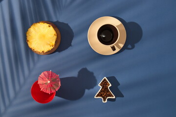 Coffee break with pineapple, drink with parasol, and with palm leaves shadows on a blue background. Minimalistic scene.