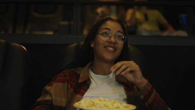 African American woman sitting in armchair watching a comedy at the cinema, eating popcorn