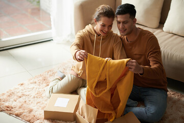 Young couple admiring the new hoody in their hands while sitting on the floor in the room, they...