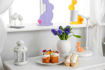 Plate with Easter cakes, holder with eggs and beautiful flowers on table near light wall
