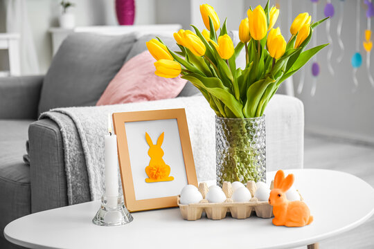 Holder with Easter eggs, photo frame, vase with tulips and burning candle in living room