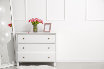 Vase with tulips and blank photo frame on chest of drawers near white wall in room