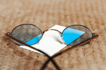 on the beige woolen surface of the soft plaid are sunglasses with gray lenses and a sheet of paper next to it for inserting text; the theme of vacation, tourism, ophthalmology and optics