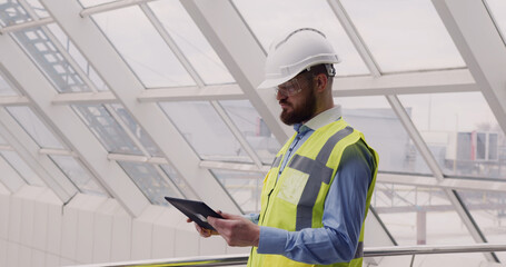Close up of male chief engineer in hardhat and vest standing inside commercial building construction site using tablet to look at architectural plan of panoramic glass roof.
