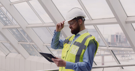 Close up of male chief engineer in hardhat and vest standing inside commercial building construction site using tablet to look at architectural plan of panoramic glass roof.