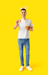Happy young man in bunny ears holding basket with Easter eggs on yellow background