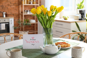 Teapot, cups, tulips and greeting card with text HAPPY WOMEN'S DAY on dining table in stylish kitchen