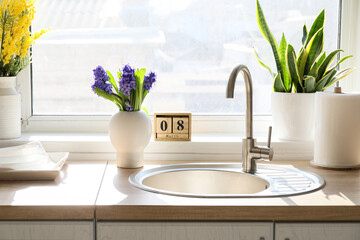 Vase with flowers near sink on kitchen counter and wooden cube calendar with date 8 MARCH. International Women's Day celebration