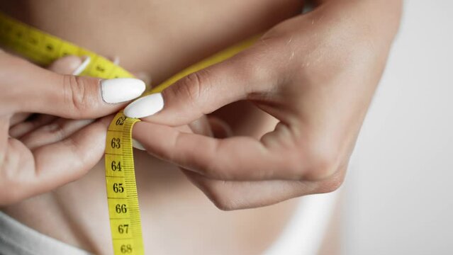 A girl is measuring her waist with a centimeter tape. The woman is taking waist scale tape and showing her. Concept of losing weight ideal figure slim stomach and proper nutrition. Close up.