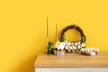 Easter cake, holder with eggs and beautiful wreath on table near color wall