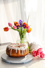 Tasty Easter cake and vase with flowers on table in room
