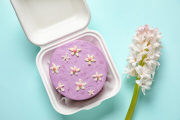 Plastic lunch box with tasty bento cake and flowers for International Women's Day celebration on turquoise background