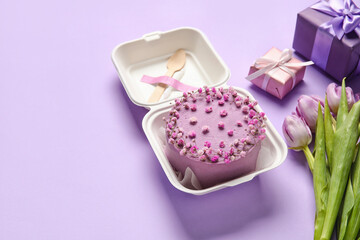 Plastic lunch box with tasty bento cake and flowers on purple background