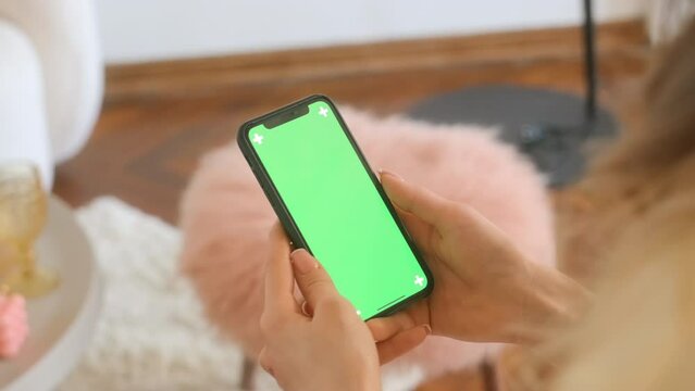 Young woman sitting on sofa at home and uses smartphone with green screen Mock-up. 4k