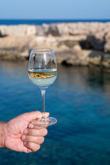 Man's hand holding glass of white dry white wine with view on rocks and blue sea bay water near Protaras touristic town on Cyprus